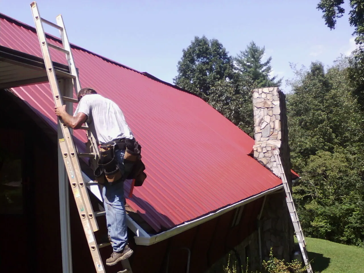 Roofer on a ladder beside a red metal roof and stone chimney — Soffit & Fascia Repair by Roofing Brothers in Summerville